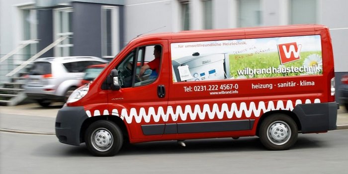 Ein roter Transporter mit einem bunten Werbeaufdruck fährt schnell vorbei, während andere Autos im Hintergrund stehen.