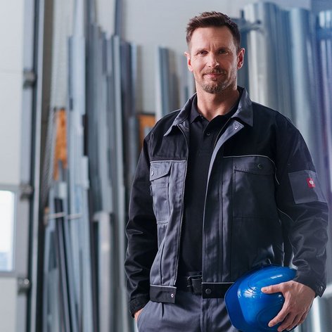 In einer hellen Werkstatt posiert ein Mann in schwarzer Arbeitskleidung mit einem blauen Helm, der bereit für die Arbeit aussieht.