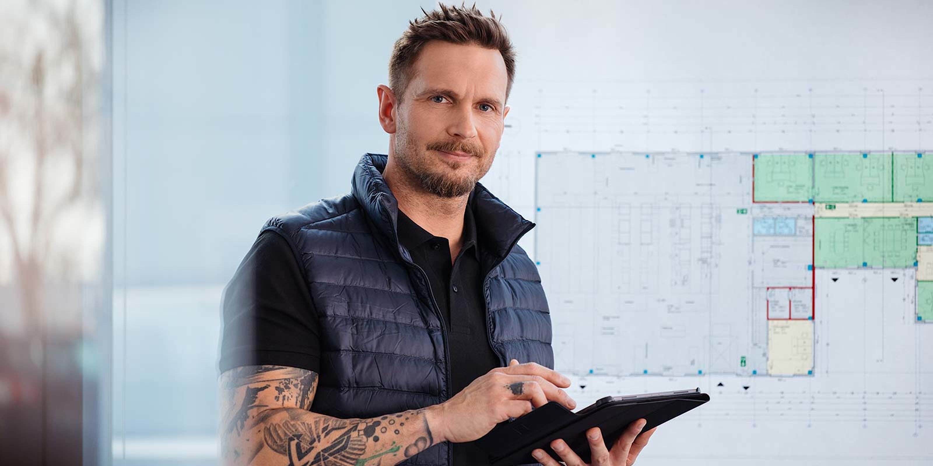 Ein Mann mit Tattoos hält ein Tablet und lächelt, während er vor einem großen Bauplan steht, der verschiedene Räume zeigt.