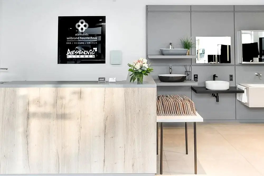 Ein heller Empfangsbereich mit einer Holztheke, einer Blumenarrangement und einer modernen Badezimmerausstellung an der Wand.