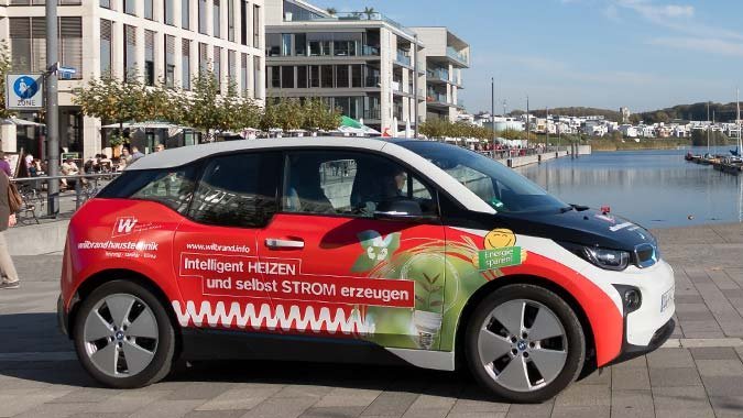 In der Nähe eines Hafens steht ein werbewirksames Elektroauto, während Passanten die Aussicht auf das Wasser genießen.